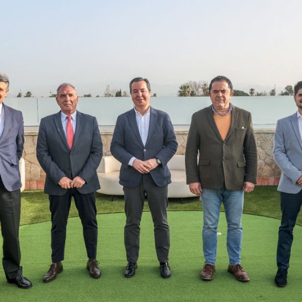 Cinco hombres de pie posan formalmente en una terraza exterior con césped artificial y mobiliario, bajo el sol de un día claro.
