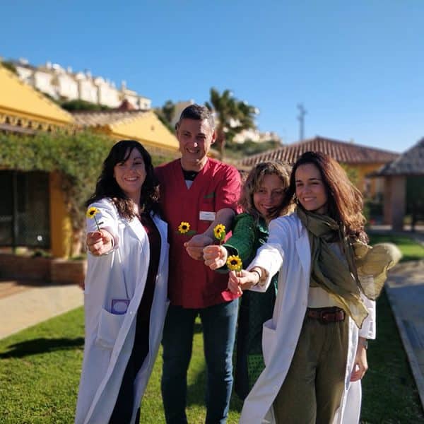 Cuatro profesionales sonrientes (tres mujeres, un hombre) en batas y uniforme, extienden pequeñas flores amarillas al espectador, en un jardín soleado con edificios amarillos de fondo.