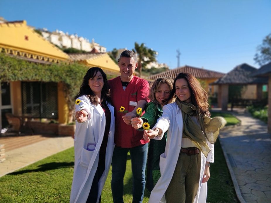 Cuatro profesionales sonrientes (tres mujeres, un hombre) en batas y uniforme, extienden pequeñas flores amarillas al espectador, en un jardín soleado con edificios amarillos de fondo.