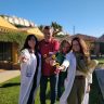 Cuatro profesionales sonrientes (tres mujeres, un hombre) en batas y uniforme, extienden pequeñas flores amarillas al espectador, en un jardín soleado con edificios amarillos de fondo.