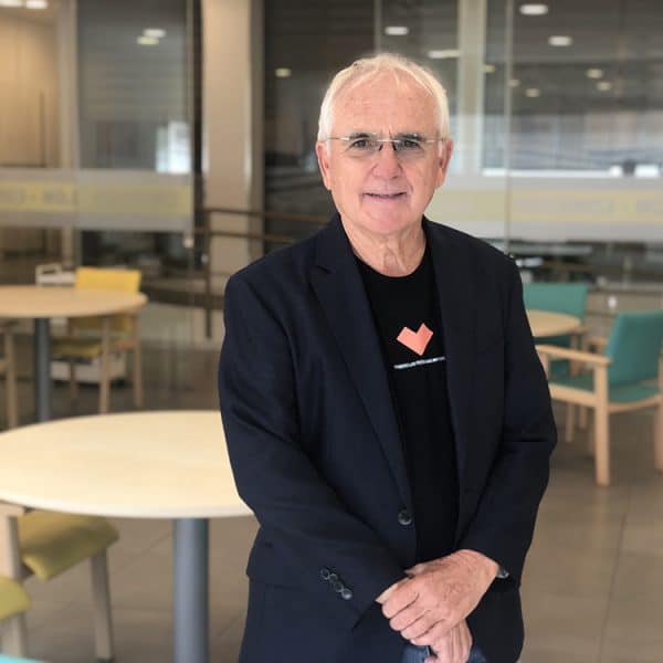 Hombre de pelo blanco y gafas mira a cámara. Viste blazer azul sobre camiseta negra con corazón naranja y el texto "THERAPEUTIC PERCEPTION". Fondo de interior moderno con mesas y sillas.