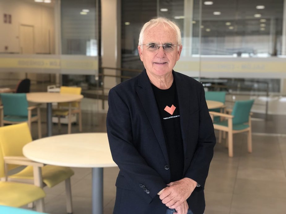 Hombre de pelo blanco y gafas mira a cámara. Viste blazer azul sobre camiseta negra con corazón naranja y el texto "THERAPEUTIC PERCEPTION". Fondo de interior moderno con mesas y sillas.