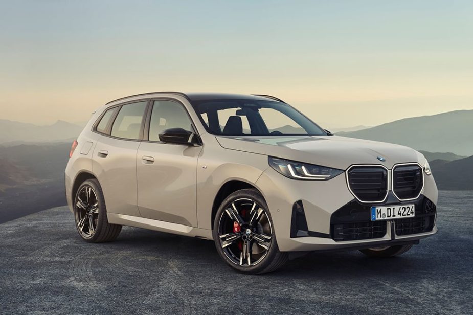 Un SUV BMW beige, modelo X1, estacionado en un camino asfaltado, visto desde un ángulo frontal-derecho. Al fondo, un cielo claro y colinas difusas.