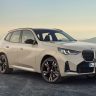 Un SUV BMW beige, modelo X1, estacionado en un camino asfaltado, visto desde un ángulo frontal-derecho. Al fondo, un cielo claro y colinas difusas.