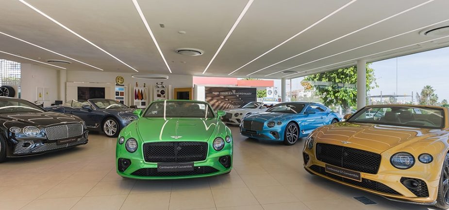 Varios coches deportivos de lujo, encabezados por un Bentley verde, están expuestos en un brillante concesionario interior. El suelo es pulido y el techo tiene iluminación de tiras.