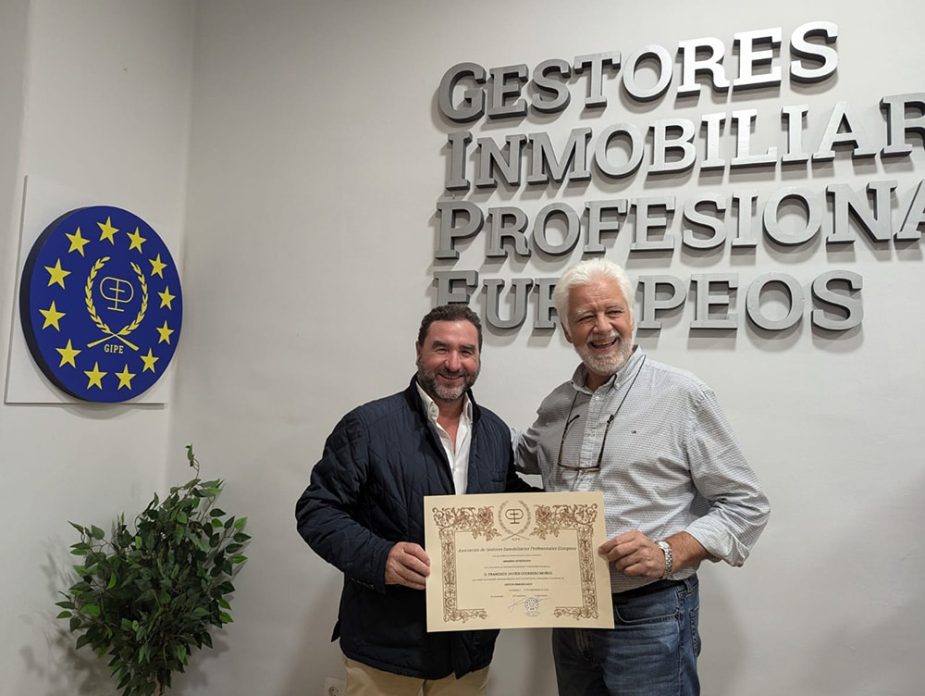 Dos hombres sonrientes sostienen un certificado enmarcado. El contexto es una oficina con el letrero en la pared: GESTORES INMOBILIARIOS PROFESIONALES EUROPEOS.