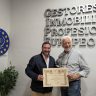 Dos hombres sonrientes sostienen un certificado enmarcado. El contexto es una oficina con el letrero en la pared: GESTORES INMOBILIARIOS PROFESIONALES EUROPEOS.