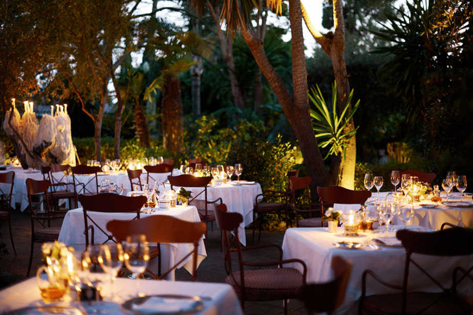 Mesas de comedor blancas con vajilla y velas encendidas, listas para cenar en un jardín exterior, rodeadas de vegetación exuberante bajo iluminación cálida.