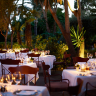 Mesas de comedor blancas con vajilla y velas encendidas, listas para cenar en un jardín exterior, rodeadas de vegetación exuberante bajo iluminación cálida.