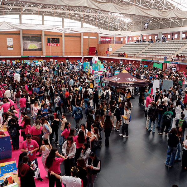 Multitud de personas se congregan y caminan en un gran pabellón interior, rodeadas de puestos y con gradas visibles al fondo, simulando un evento o feria.