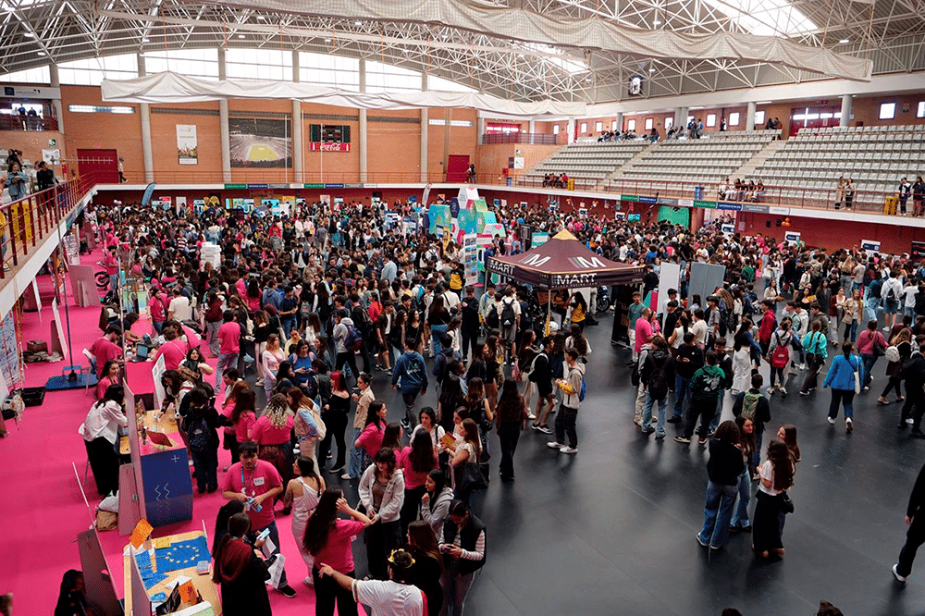 Multitud de personas se congregan y caminan en un gran pabellón interior, rodeadas de puestos y con gradas visibles al fondo, simulando un evento o feria.