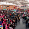 Multitud de personas se congregan y caminan en un gran pabellón interior, rodeadas de puestos y con gradas visibles al fondo, simulando un evento o feria.