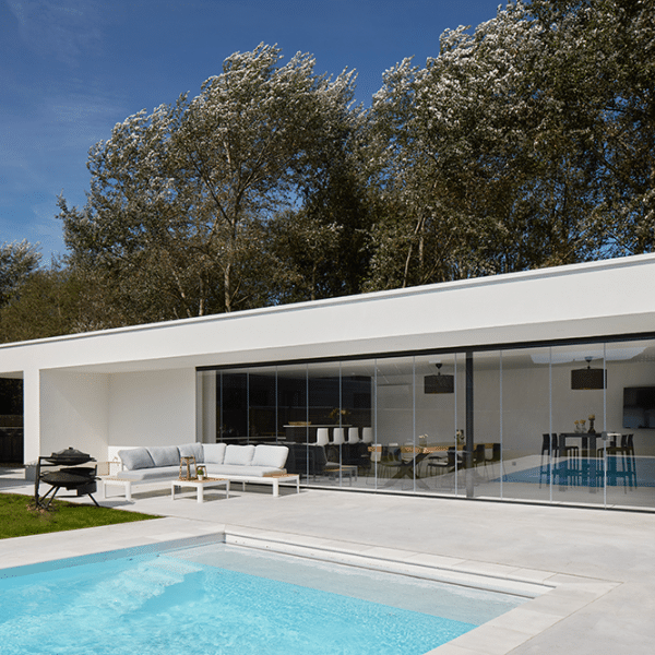 Edificio moderno blanco con amplios ventanales conecta un salón interior con un patio amueblado y una piscina, rodeado de árboles bajo un cielo azul.