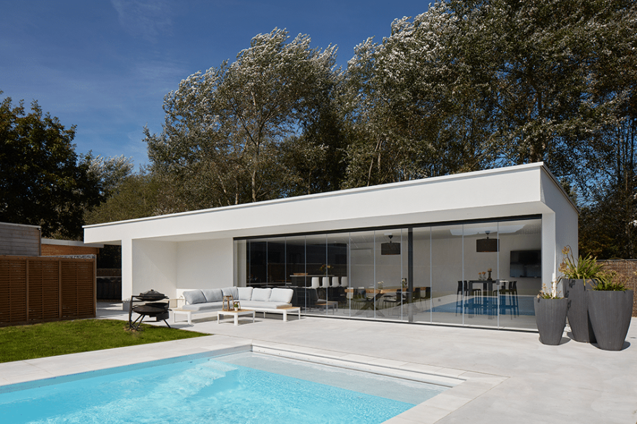 Edificio moderno blanco con amplios ventanales conecta un salón interior con un patio amueblado y una piscina, rodeado de árboles bajo un cielo azul.