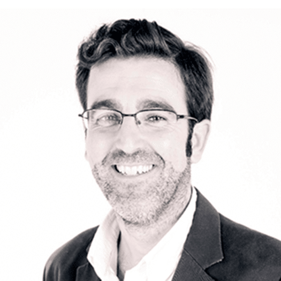 Hombre con gafas y barba, sonriendo con dientes visibles, en un retrato de estudio en blanco y negro sobre fondo blanco.